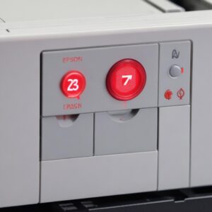 A close-up shot of an Epson L3210 printer's control panel, specifically highlighting the blinking red ink and paper indicator lights. The background is slightly blurred.