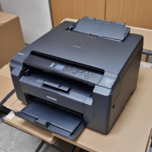 Seorang pria muda sedang memeriksa kondisi fisik Printer Epson L3210 bekas, memegang bagian bodi printer dan melihat detailnya dengan serius. Latar belakang toko elektronik bekas yang rapi.